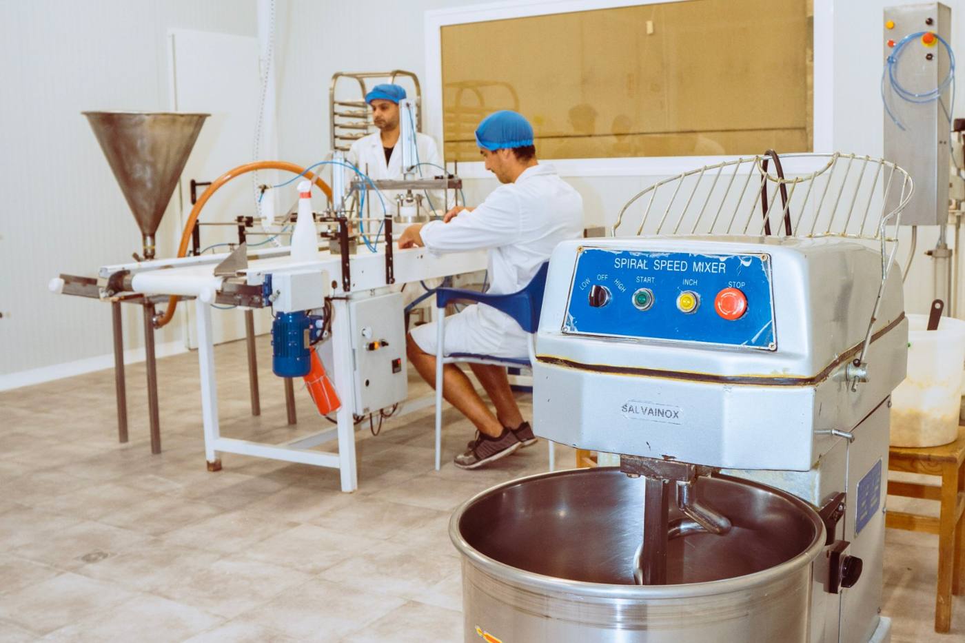Dos operarios con bata y gorro trabajando en la línea de producción del obrador, con una amasadora en primer plano.